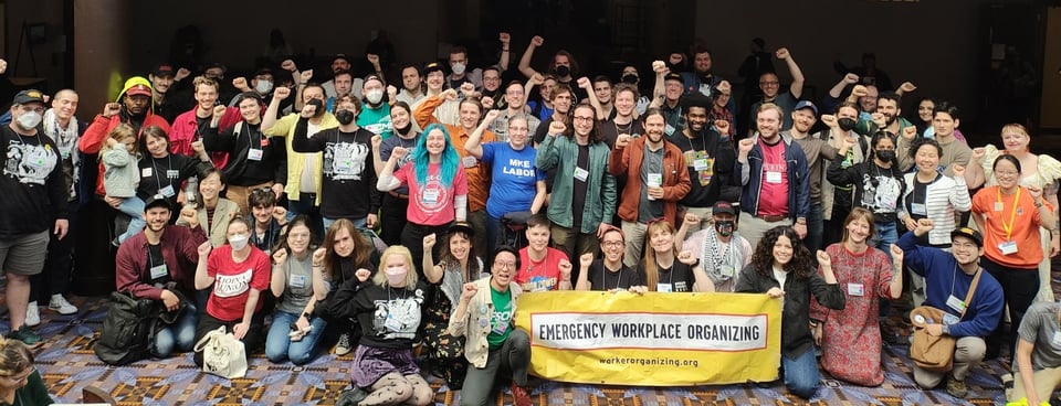 About 60 members of the Emergency Workplace Organizing Committee standing or kneeling with a fist raised at Labor Notes 2023.