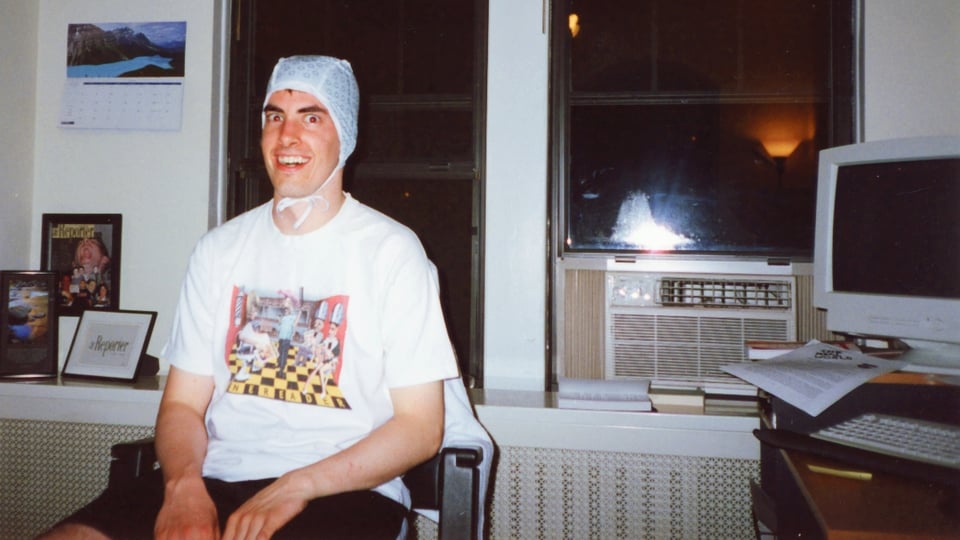 A smiling person wearing a plastic cap, sitting in front of a window with an a/c unit, and a large square computer monitor on a desk to the right