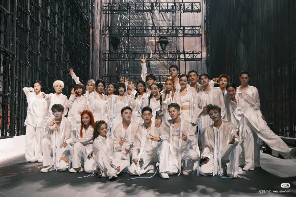 A large group of dancers in white costumes with translucent gauze tops, white tassels, and lightly smeared pale blue and white makeup, posing for the camera. They look like they're backstage wedged between a lattice scrim and a metal grid wall, with lighting scaffolding in the rear.