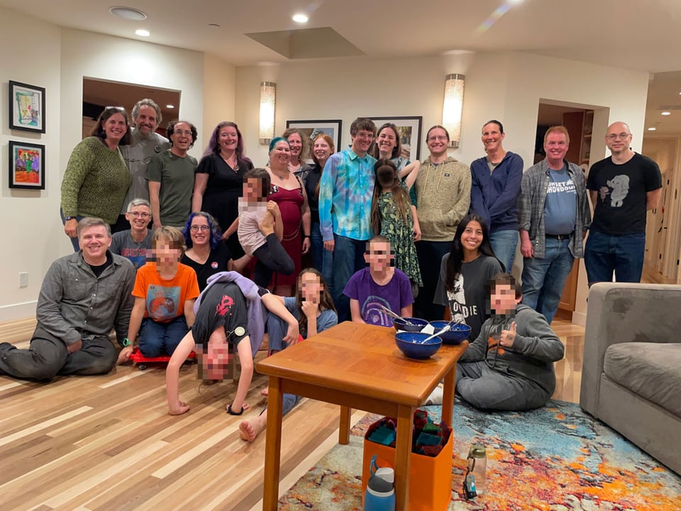 A large group of people including children stand and sit together to have a group shot. The childrens' faces are blurred.