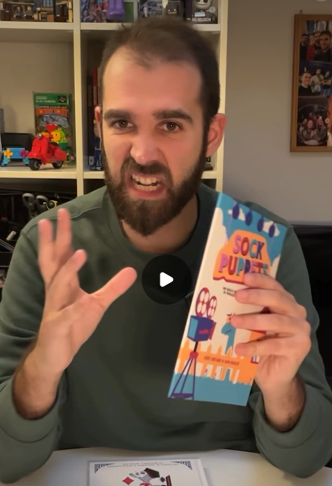 A man gestures with his right hand as he holds Sock Puppets in his right hand.
