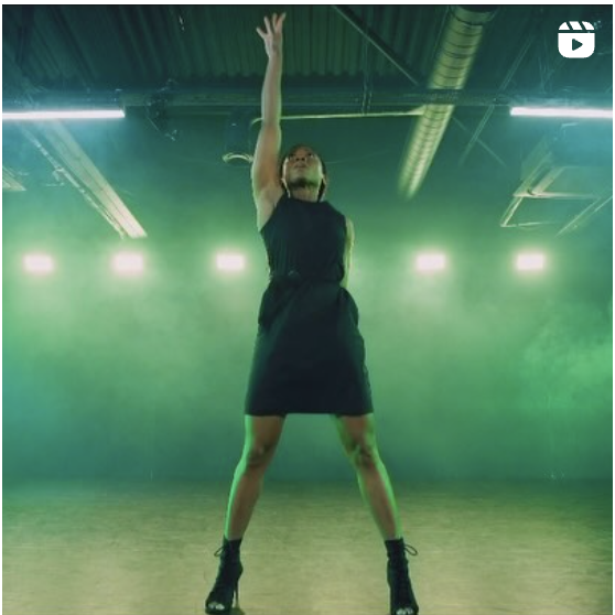 Still of me in a black dress and heels with an arm stretched out to the ceiling and I'm looking up towards my hand. The lighting is green tinted and there's smoke in the background.