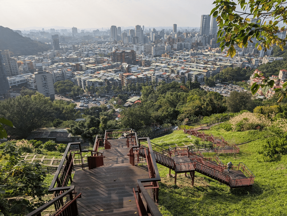 An outdoor shot from the side of a hill. Below is a long series of steps down the green hill. In the background is Taipei's skyline, which closest is a mass of cheap-looking five-story buildings then, further back is the skyscrapers and modernity.