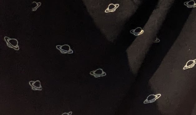 Foto de un estampado de una camiseta: la tela es negra y hay planetitas con su anillo flotando.
