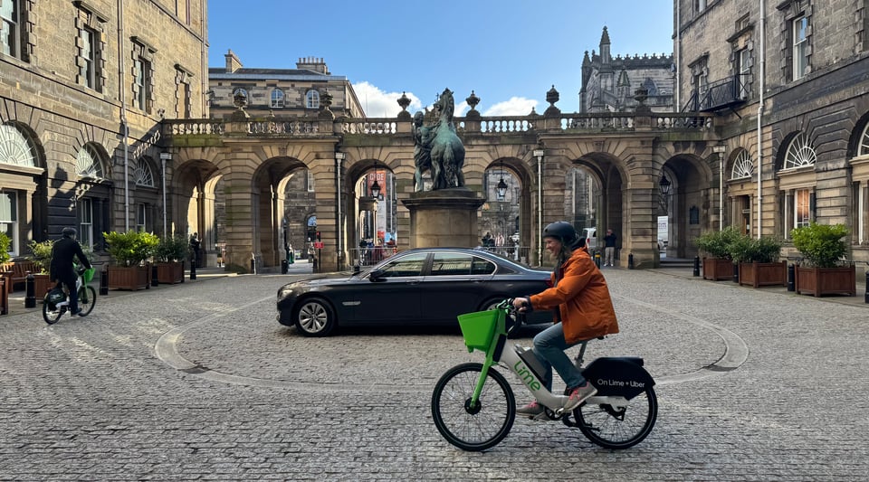 Two councillors trial white and green ebikes from provider lime round a historic courtyard on a bright and fresh spring day. Someone's left their motor in the middle. Can't park there mate.