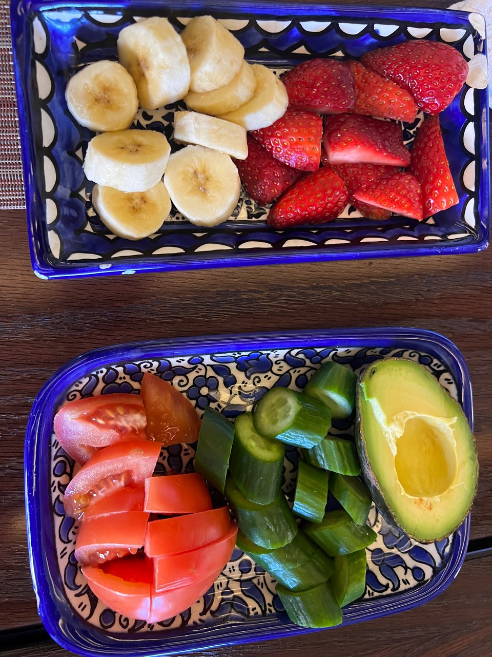 A blue and white ceramic dish holding sliced banana and strawberries; another blue and white ceramic dish holding sliced tomatoes, cucumber and half an avocado
