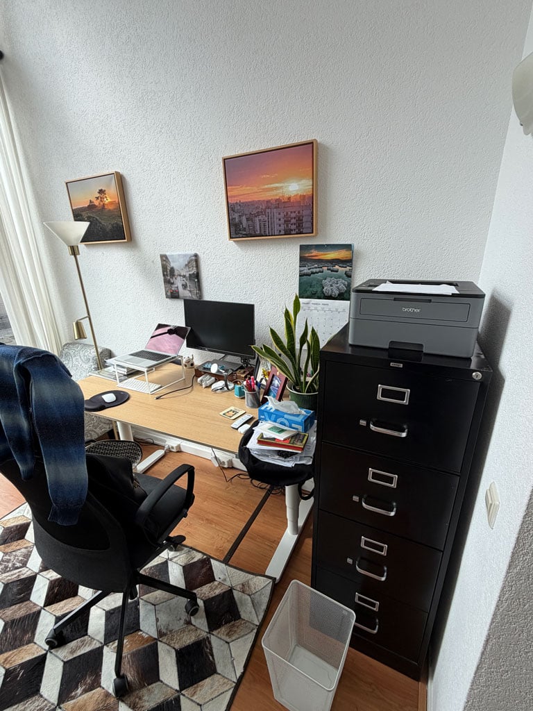 A home office with a desk, an office chair and a black filing cabinet. The desk is covered with a computer, monitor and some papers and supplies.