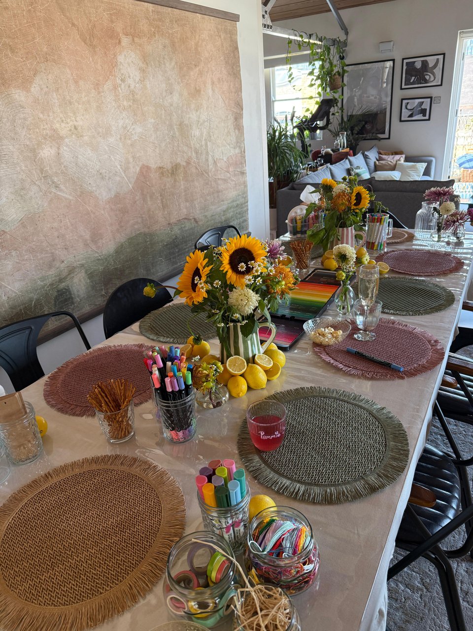 A beautifully arranged table set up for a creative gathering or craft session, with woven placemats, jars of markers, washi tape, thread, sunflowers in vases, and decorative lemons.