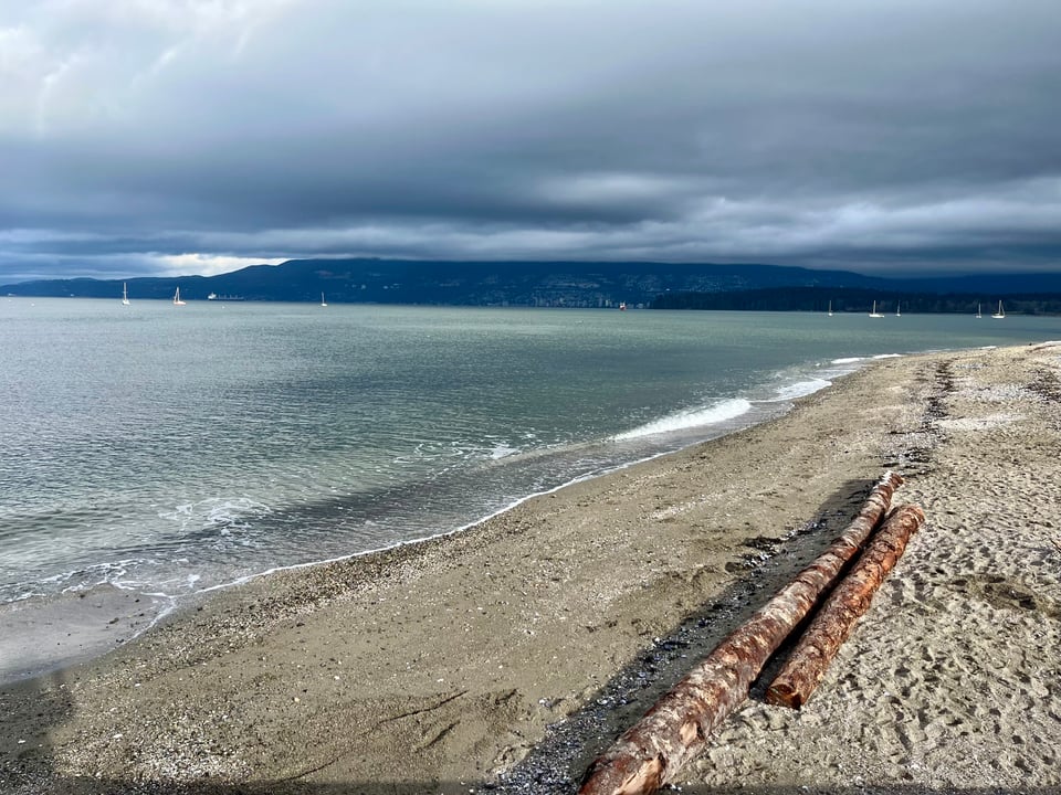 A sandy beach with bluish-green waves lapping at the shore. There are grey clouds on the head, and land on the horizon.