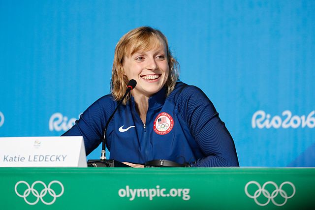 A white woman sitting at a desk with a microphone in front of her face. She's looking to the right and smiling. The table is green and has "olympic.org" and the Olympic rings printed on it.