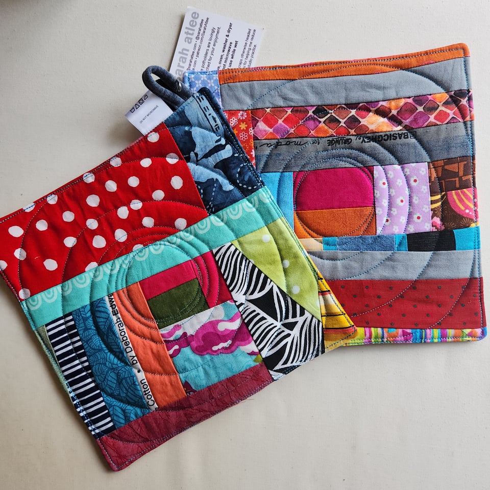 A pair of Sarah’s quilted potholders, using fabrics in all different colors, against a white fabric background.