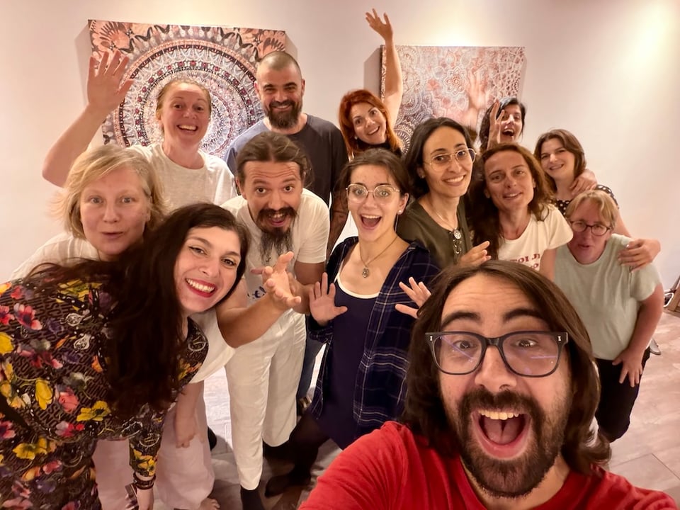 A diverse group of people is gathered together, smiling and posing playfully for a selfie in a brightly lit room with artistic wall decorations.