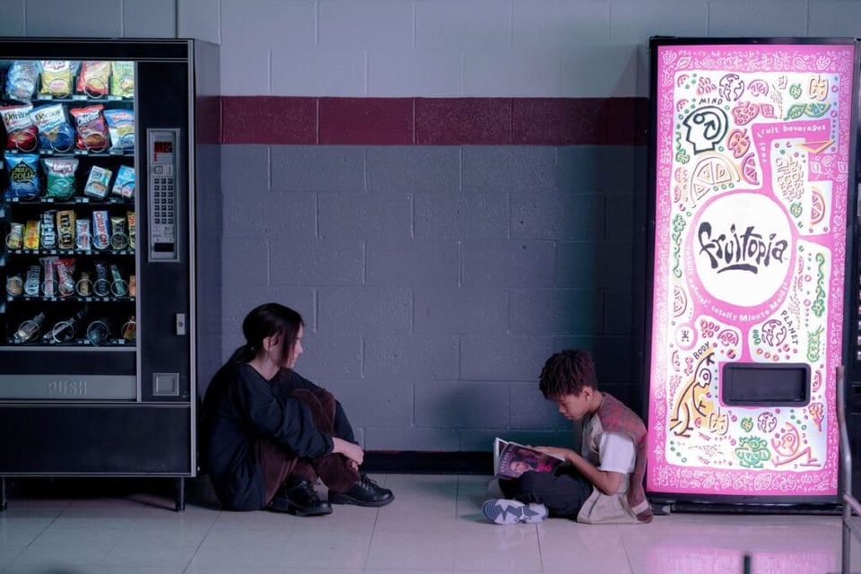 Two young people sit on the floor, facing each other between two vending machines. The person on the left looks at the person on the right, who is reading a book.
