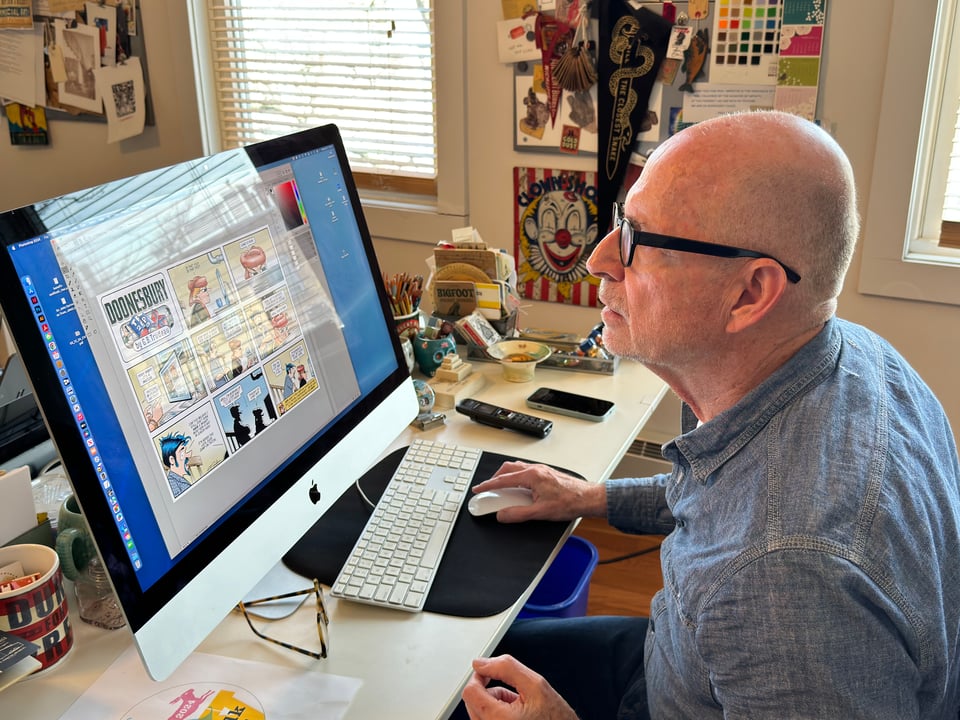 George Corsillo in a blue collared shirt sitting at a computer looking at a Sunday Doonesbury on screen in his office