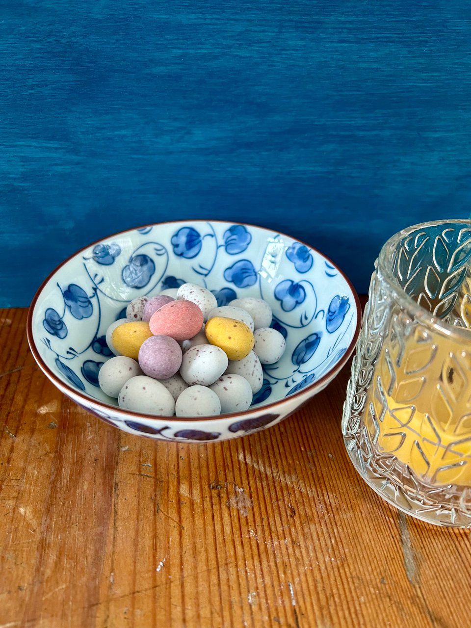 A Japanese porcelain bowl painted with blue cherry blossom on white is filled with chocolate mini eggs. They're lightly speckled and in white, pink, yellow and purple. The bowl is on an old pine trunk and there is a chunky beeswax candle in textured glass holder on the right. The background is a vibrant teal.