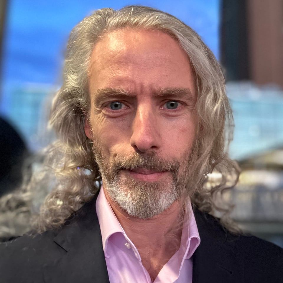 Close-up photo of a white man in a navy suit jacked and pink button-down shirt. He has long hair that is being tousled by the wind and his eyes betray how exhausted he is