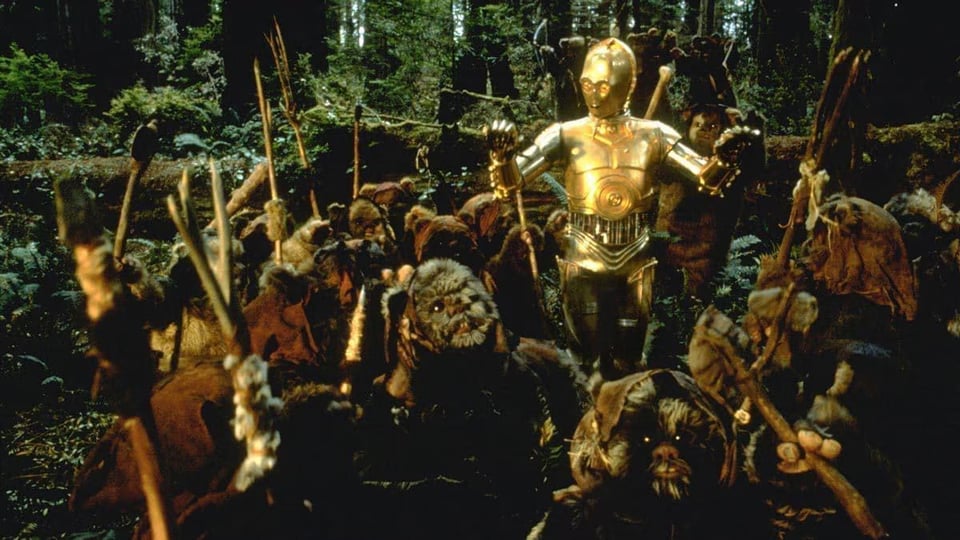 A still from Star Wars: Return of the Jedi, in which C-3PO is hoisted up by the cuddly Ewoks, who believe he's a god