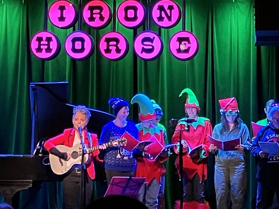 A person in a red tuxedo jacket with black piping plays an acoustic guitar accompanied by people in elf costumes reading from hymnals. They are in front of a green-lit curtain and underneath a red-lit sign reading I-R-O-N H-O-R-S-E