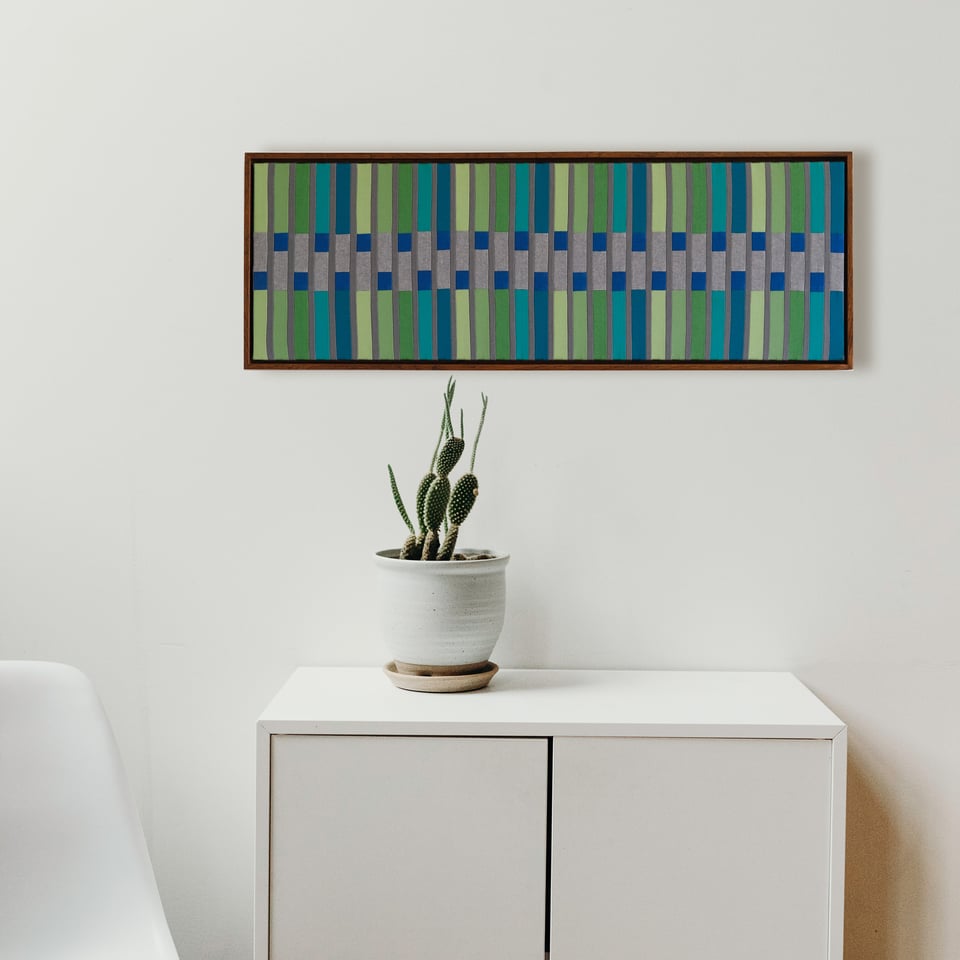 A linear, geometric artwork, made with fabrics, on the wall of a white room. The artwork is green and blue.