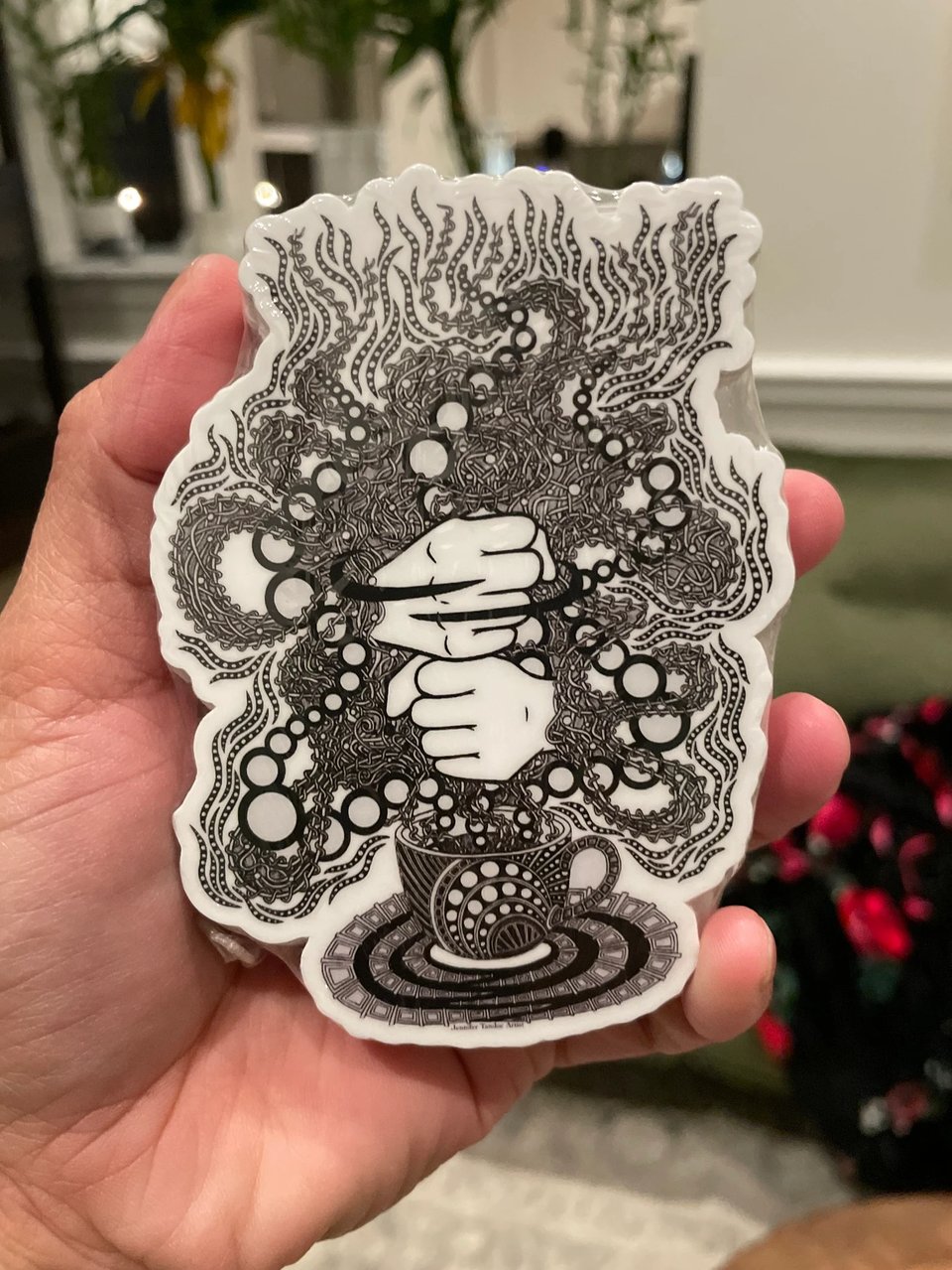Hand holding a black and white sticker, depicting a cup of coffee spilling a cloud of pixellated psycedellic steam. Hands superimposed over the cloud sign the ASL word COFFEE.
