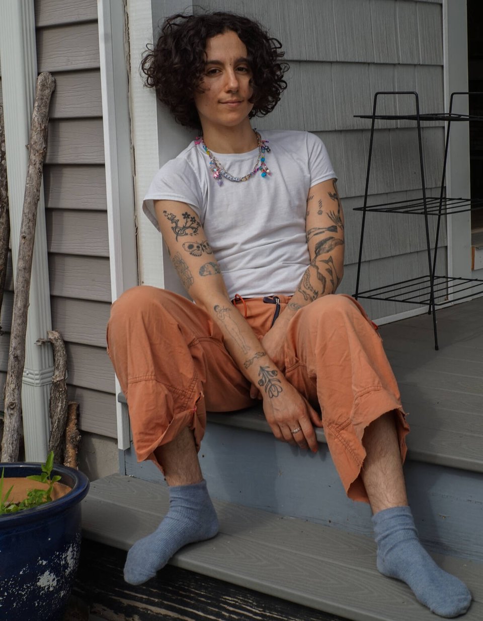 A person sits on the top step on a porch outside a house, wearing flowy orange pants, a tight white t-shirt and gray socks. The person has a mop of curly hair, and their arms feature tattoos.