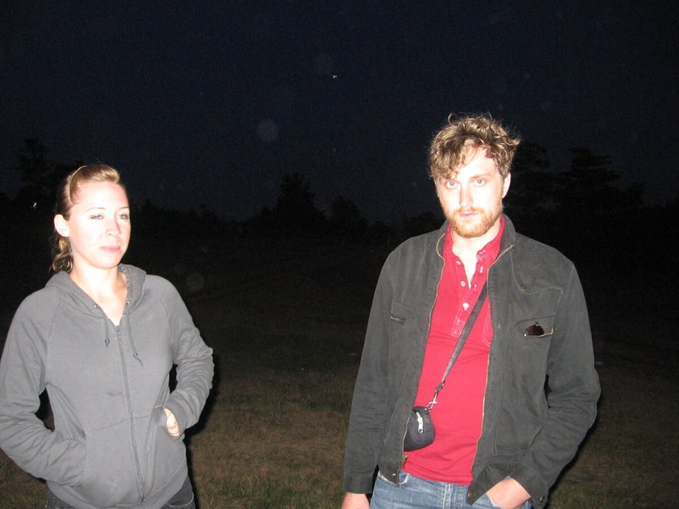 A woman with her hair up with her hands in the pockets of a gray hoodie stands next to a man wearing a trucker-style jacket and a red polo shirt at night, standing on grass.