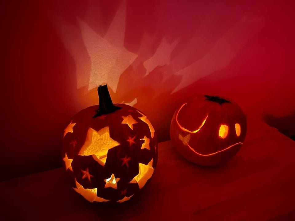 A pair of jack-o-lanterns light up a dark room. One is carved like the character Haro from Gundam with a cute face, and the other has stars cut and etched out all over. Star-shaped lights dance on the wall behind the pumpkins, giving it a magical effect.