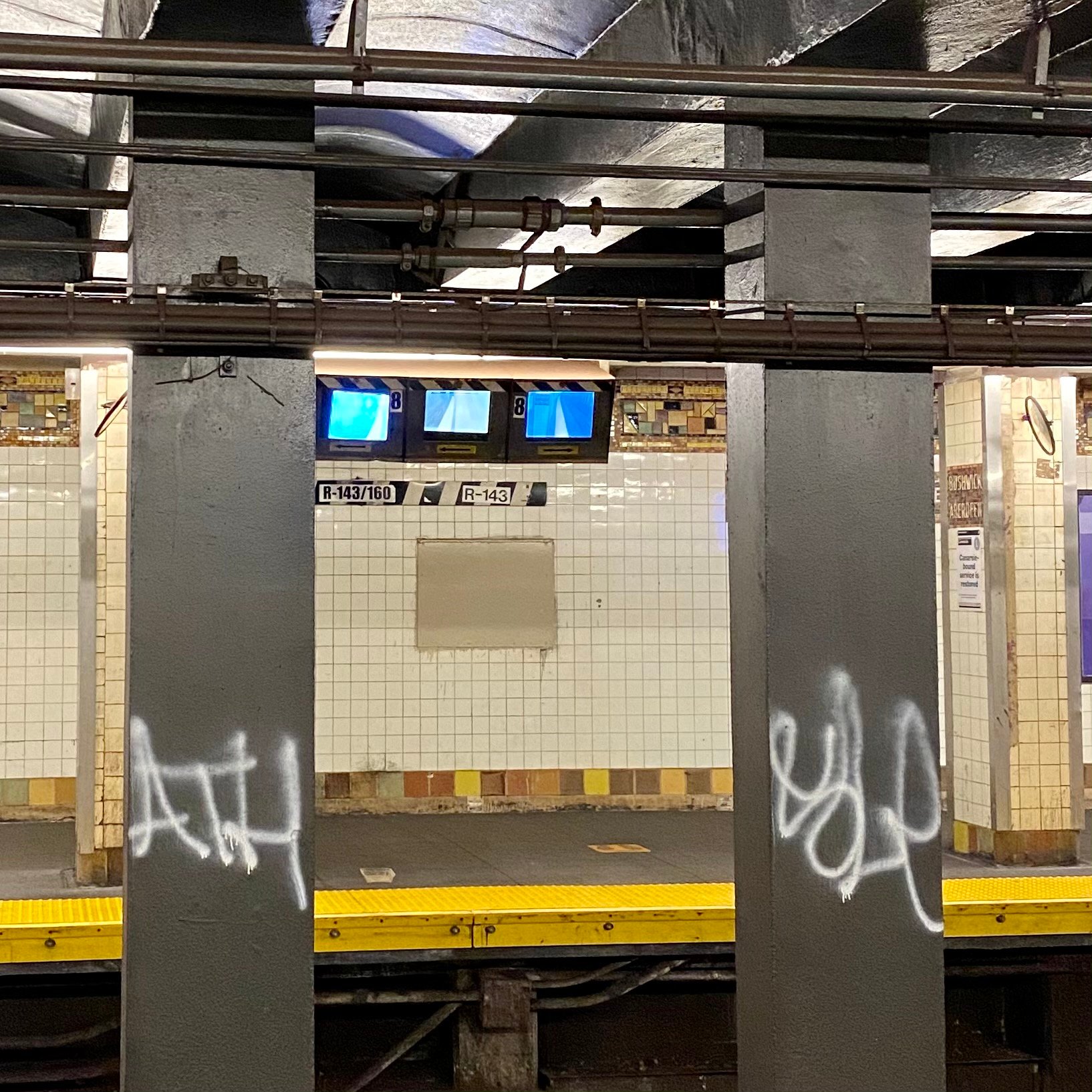 Image of the track on an MTA metro station. Between two gray pillars, there are three small TV's hanging from the ceiling, showing the incoming activity on the train tracks.