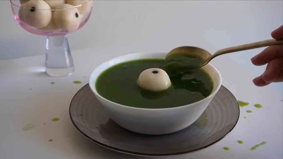 A spoon scoops up some green soup. A dumpling shaped like an eyeball floats in the middle of the bowl.