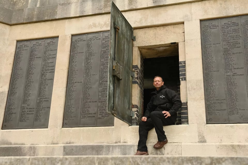 A ashlar stone memorial wall beneath a tower. The wall has huge bronze plaques on them with the names of the dead. One of the plaques has been hinged outwards to reveal a doorway where a white man in dark clothes is sitting.