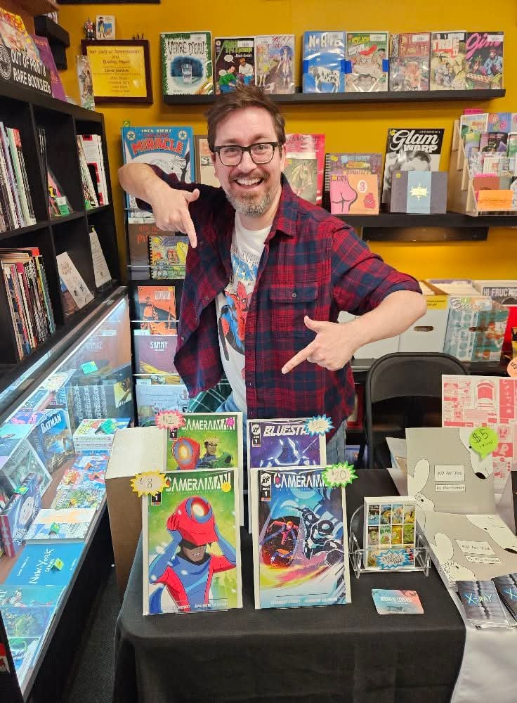 A nerdy man points at comic books he wrote from a small table in a comic book store.