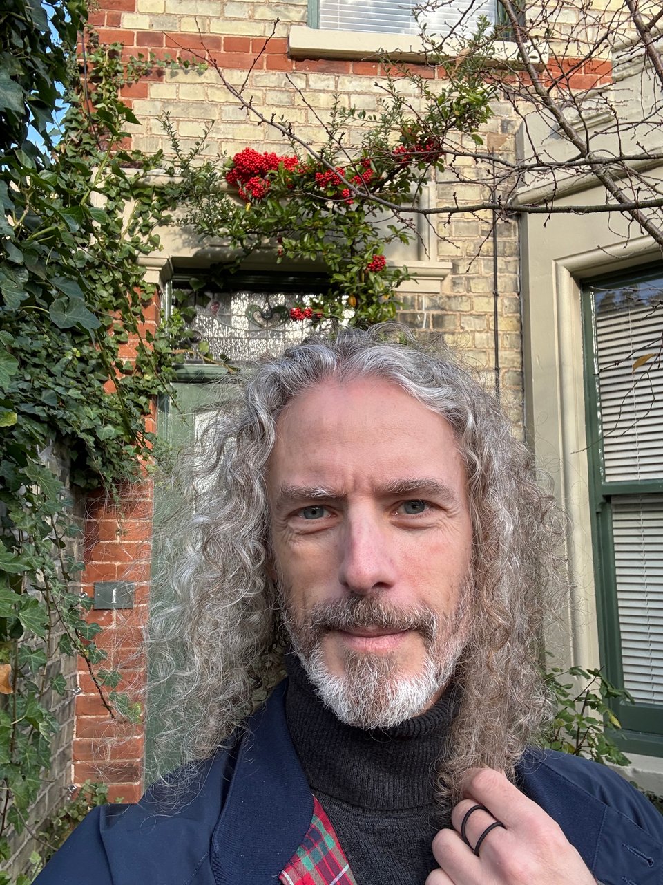 Selfie of white man with long curly hair standing in front of a flat in Cambridge. There's ivy & holly climbing up around the front door.