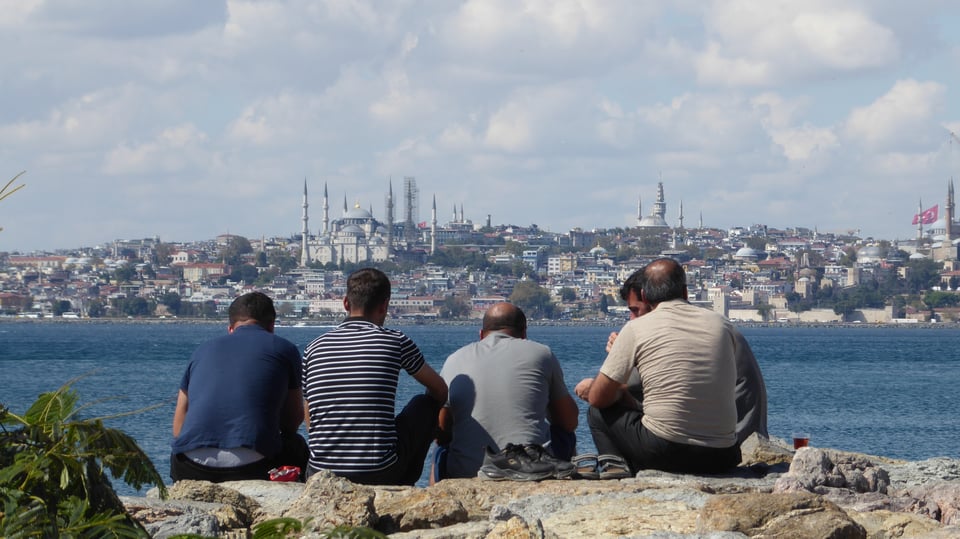 Photograph of a group of men sitting on a sea wall with their backs to the camera, looking across a body of water. On the other side of the water, in the distance, are the dome and minarets of a large mosque.