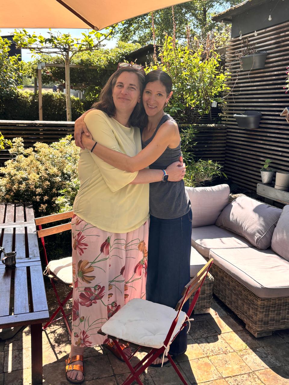 Two women stand in a patio of a front garden hugging looking at the camera.