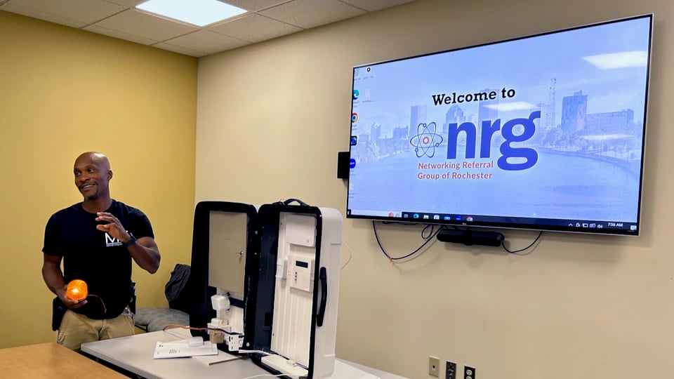 Shantae has a demo of a home alarm system inside an open hardshell suitcase. He stands next to it and in front of the NRG Welcome TV screen.