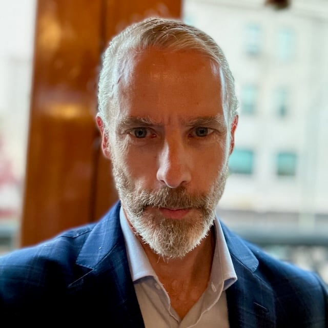 Photo of white man in light blue dress shirt and navy blazer, in gold-ish lighting