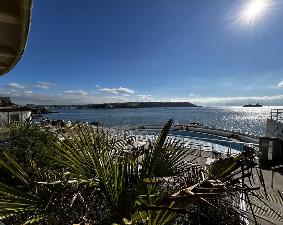 A bold blue sky reflected in the sea. The cruise ship is silhouetted in the Sound. In the foreground are a range of subtropical plants, almost hiding the sun terraces seating and sun loungers. The empty pool is just visible.