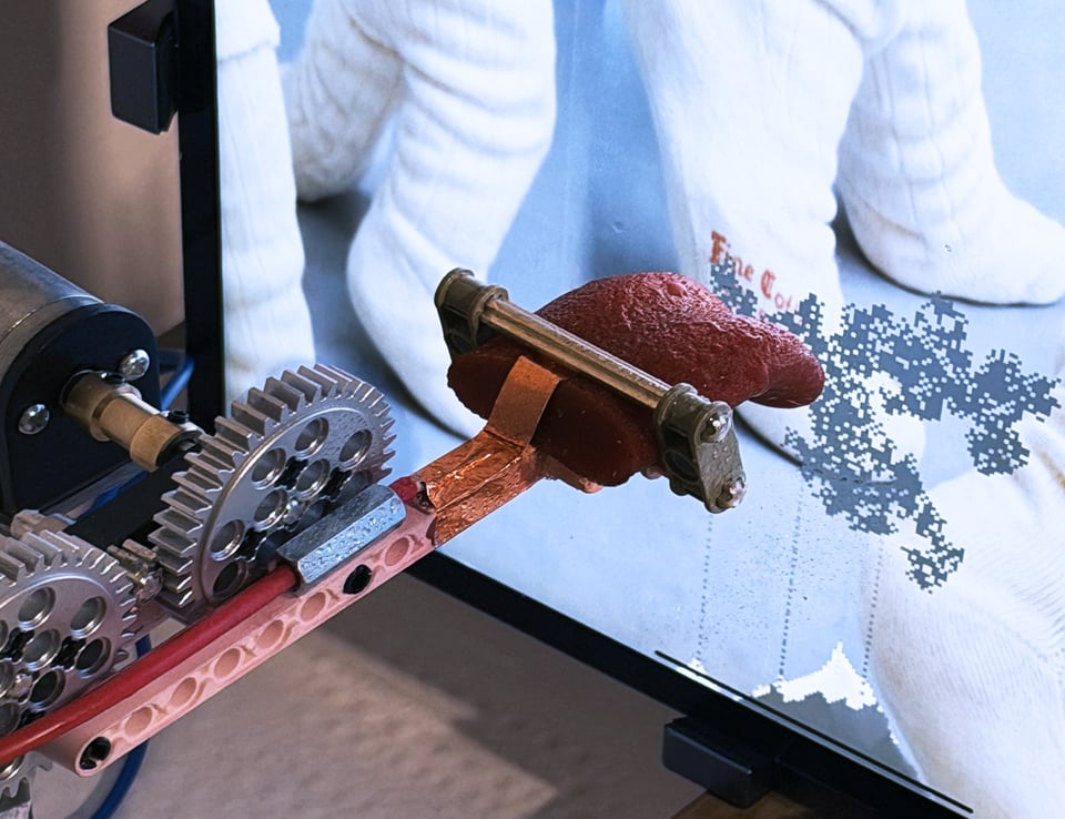 A silicone tongue mounted to a machine, with prominent metal gears, licking a screen. The screen has a pale blue and white image of socks, which are turning into pixelated animation on the screen - like fungus and falling sand - where the tongue touched it
