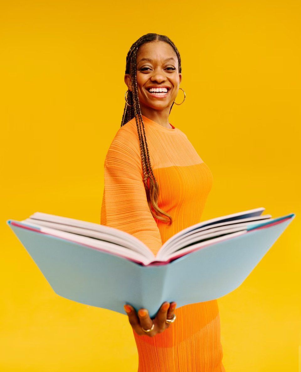 I'm captured in a joyful moment, dressed in a bright orange long-sleeve dress, holding a large open book towards the camera. My hair is styled in long braids, and I'm smiling broadly against a cheerful yellow background. The scene exudes a warm and inviting atmosphere, emphasizing my enthusiasm for reading.