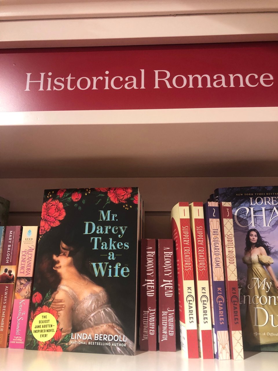 a photo of a bunch of books on a shelf below a sign reading "Historical Romance." in the center of the photo are the red spines of two copies of A BLOOMY HEAD, by J. Winifred Butterworth. to the left is Mr. Darcy Takes a Wife by Linda Berdoll and to the right are the Will Darling books by KJ Charles.