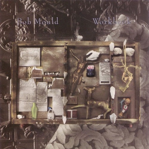 A photograph of a frame with a number of objects placed within. There's too many things to describe, and I'm not sure what all of them are, but I see some shells, a padlock, a figurine of a deer, some clippings, another figurine of a smiling man waving his arm. Behind the frame are some patterns/textures, one looks like it's made of metal and another is a pattern of leaves that looks like it might be from a couch. The whole image is very light grey and light brown.