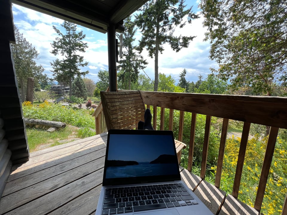 POV photo of sitting on a cabin porch with feet up, laptop on legs. Trees are all around and the sky is bright. On the laptop display is a photo of some islands.