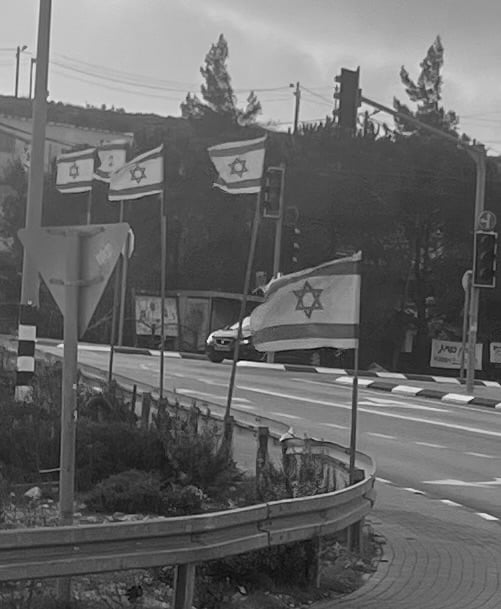A black and white image of a paved road, with five Israeli flags on flagpoles on the side of the road