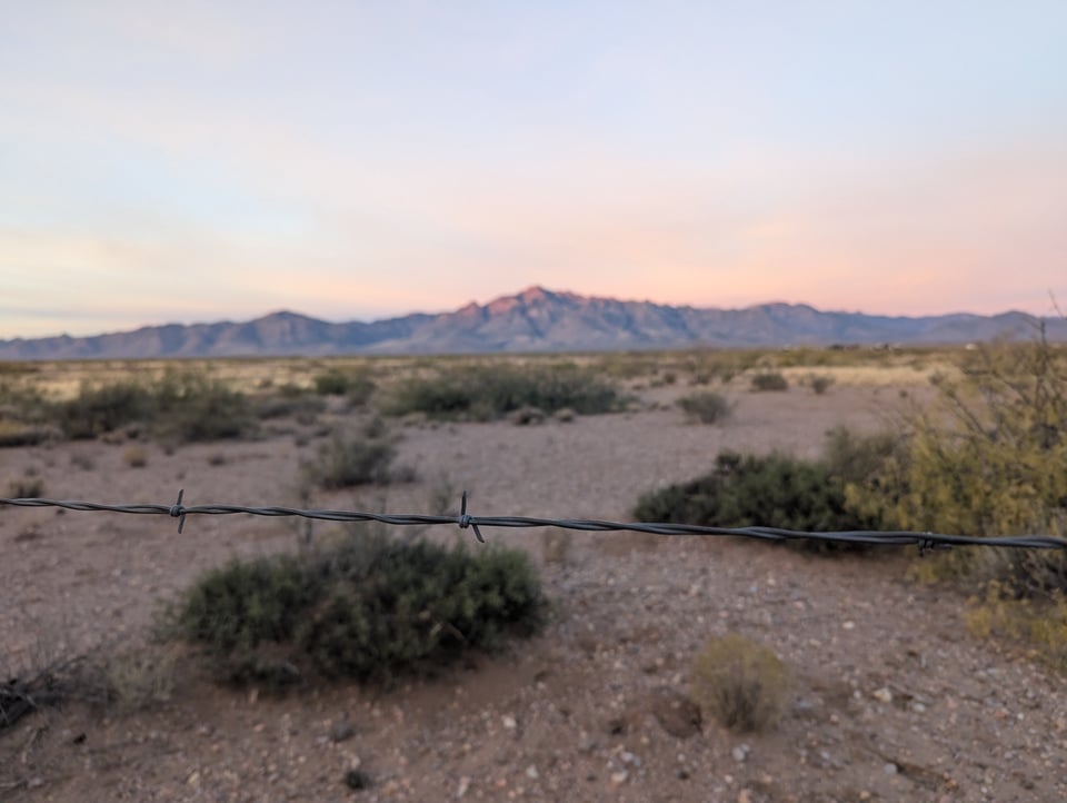 A close-up shot of a length of barbed wire stretched across a landscape dotted with sagebrush. That same mountain is in the background, blurred and out of focus.