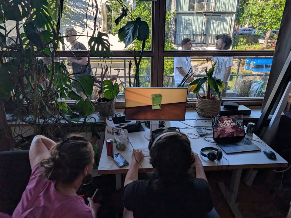 Two people play a video on a computer monitor placed in front of a window. Beyond the window is sun, green, an Amsterdam canal, and a few folks chatting in groups.
