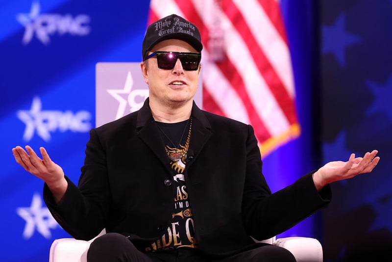 Elon Musk at CPAC 2025, dressed like a nightclub bouncer at a crypto rally, shrugging as if to say “What, me destabilize democracy?”