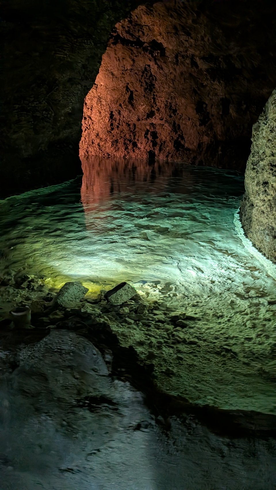 A pool of water in a stone cave, lit from beneath so it is glowing soft green. A rock wall separates the viewer from a curve in the cave, which is illuminated from behind with a red-yellow light. It looks like someone in a lantern-lit boat is about to paddle around the corner.