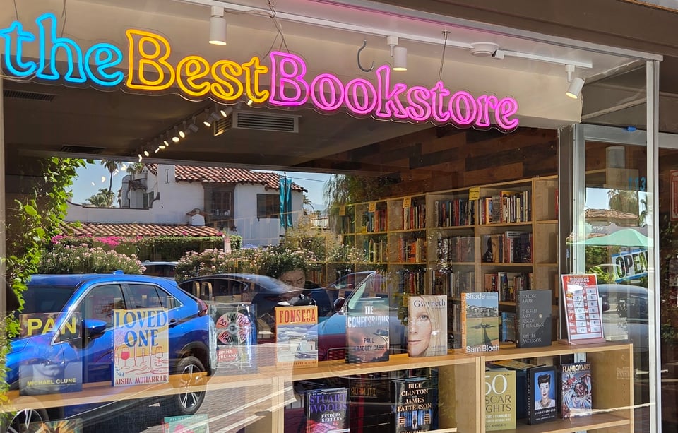 View of a bookstore from the sidewalk outside. Through the window we see shelves of books and a prominent neon sign reading "the Best Bookstore"
