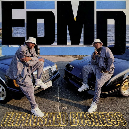 Two young Black men, dressed in identical loose-fitting grey clothing, white sneakers and white kangol hats, each sit on the hood of a car. They look at the camera. The cars are parked facing each other at an angle. Beyond them is the ocean.
