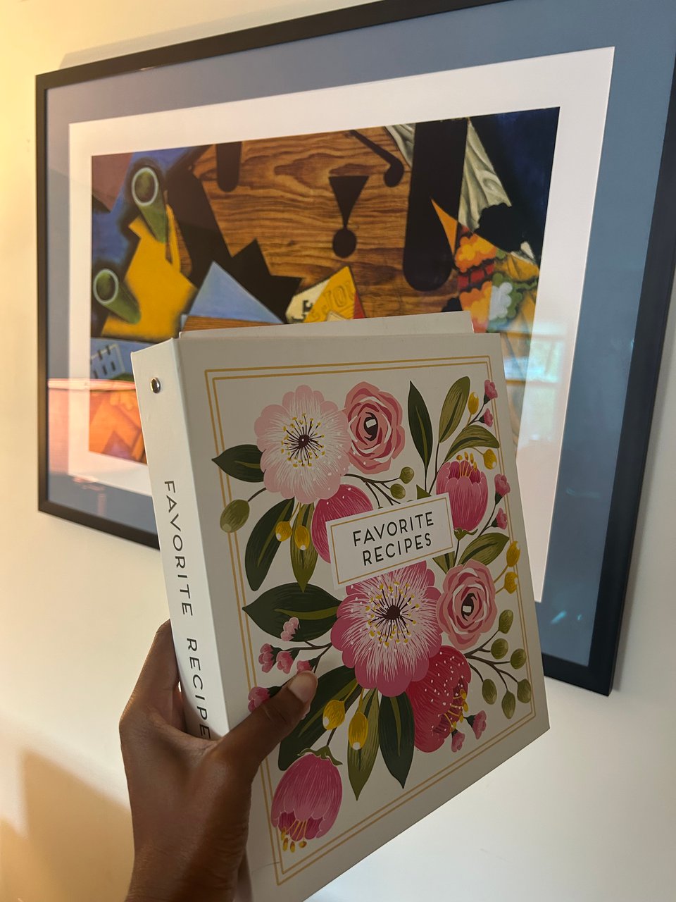 a photograph of a brown hand holding a repurposed recipe binder with pink flowers on the cover.